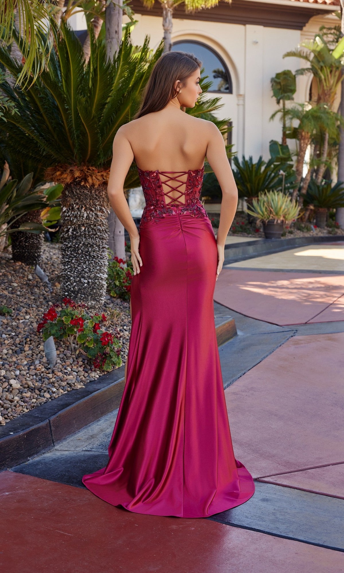 A woman stands outdoors among lush tropical plants wearing the Nox Anabel A1743, a long strapless satin prom dress with a fitted bodice, lace-up back, and high-leg slit.